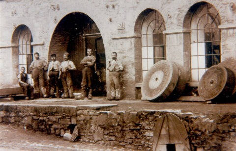 Historische Fotografie mit Arbeitern vor einem Fabrikgebäude und großen Schleifsteinen